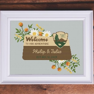 May include: Cross-stitch art in a white frame. The design features the text "Welcome to our Adventure" and "Philip & Julia EST. DEC. 12, 2025". It includes a National Park Service emblem and floral accents in orange, yellow, and white.