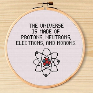 May include: A cross-stitch embroidery in a wooden hoop. The text reads "THE UNIVERSE IS MADE OF PROTONS, NEUTRONS, ELECTRONS, AND MORONS." Below the text is a black and white atom graphic with red and blue details.