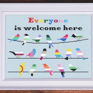 Könnte beinhalten: Gerahmtes Kreuzstich-Kunstwerk mit weißem Rahmen. Es zeigt bunte Vögel auf Drähten vor hellblauem Hintergrund. Der Text "Everyone is welcome here" ist in verschiedenen Farben über den Vögeln gestickt.