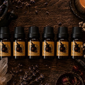 May include: Six identical amber glass essential oil bottles with black caps, each featuring a tan label with a black graphic of a cat wearing a witch hat behind a cauldron and the text Sticks and Stones NATURALS printed in black ink.