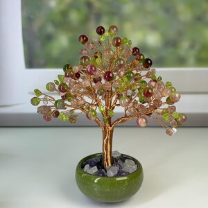 Peut inclure: Une sculpture d'arbre en fil métallique décoratif avec des branches ornées de petites perles rondes polies dans des tons de rose, vert et rouge foncé. Le tronc est en fil de cuivre torsadé et la base est un pot en céramique vert rempli de pierres transparentes et violettes.