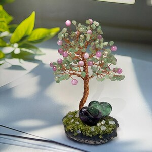 Peut inclure: Une sculpture d'arbre en fil de cuivre torsadé ornée de petits éclats de pierre vert clair et de perles roses rondes. La base est composée de deux pierres violet foncé en forme de cœur posées sur une surface texturée en mousse verte.