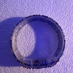 Op de afbeelding: Een heldere, cirkelvormige armband met een rij kleine, sprankelende versieringen langs de buitenrand. De armband heeft een sluiting. De achtergrond is een gestructureerd, wit oppervlak.