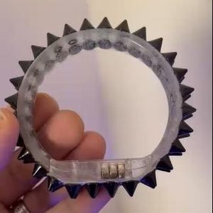 May include: A clear, round bracelet with black, pointed spikes around the outer edge. The bracelet has a clear, gel-like interior and a small, metallic clasp. The spikes are evenly spaced and create a bold, edgy look.