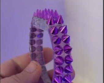 May include: A clear, spiked bracelet with metallic purple cones. The bracelet is held in a hand, showcasing the design and color. The spikes are evenly spaced around the bracelet's exterior.