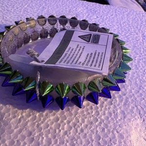 May include: Clear glass ashtray with a ring of metallic green and blue spikes around the edge. A white warning label is inside the ashtray. The ashtray is circular and sits on a white surface.