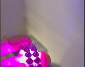 May include: A pink and silver studded bracelet. The bracelet is held in a hand and illuminated with a bright pink light. The studs are cone-shaped and evenly spaced along the length of the bracelet.