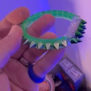 May include: A neon green bracelet with silver-colored spikes. The bracelet is made of a translucent material and is held in a person's hand. The spikes are evenly spaced around the bracelet.