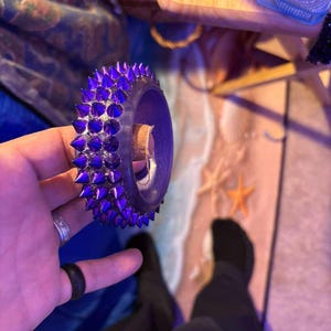 Può includere: Un braccialetto viola con punte e una fascia scura. Il braccialetto presenta numerose punte appuntite e spaziate uniformemente. Il braccialetto è tenuto su uno sfondo con una scena di spiaggia.