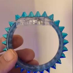 Może przedstawiać: Przezroczysta, niebiesko zabarwiona bransoletka z turkusowymi, stożkowatymi kolcami na zewnętrznej krawędzi. Bransoletka wydaje się być wykonana z przezroczystego materiału, prawdopodobnie plastiku, i jest trzymana przez dłoń.