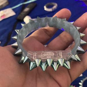 Może przedstawiać: Przezroczysta i szara bransoletka z srebrnymi kolcami. Bransoletka jest trzymana w dłoni, prezentując jej design. Kolce są równomiernie rozmieszczone wokół zewnętrznej strony bransoletki.