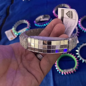 Puede incluir: Una pulsera de bola de discoteca plateada con una superficie de espejo cuadrada. La pulsera tiene una banda de arpillera y está rodeada de cristales claros y brillantes. Otras pulseras con luces de colores están en el fondo.
