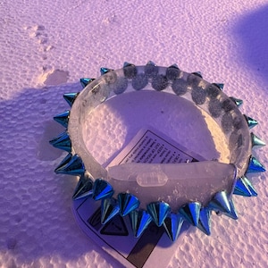 Op de afbeelding: Een doorzichtige armband met metallic blauwe spikes. De armband heeft een reeks gelijkmatig verdeelde, puntige spikes rondom de omtrek. Een label met tekst is zichtbaar.