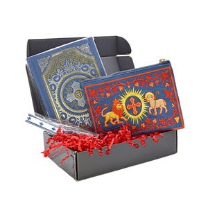 May include: A gift set in a black box, featuring a blue book with gold accents and a blue zippered pouch. The pouch has a red embroidered design with a lion, lamb, and cross. Red paper shreds fill the box.