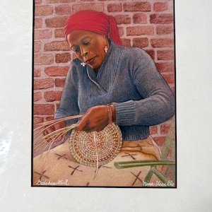 May include: A framed artwork shows a woman crafting a basket. She is wearing a red headscarf and a blue sweater. The background is a brick wall. The artwork is signed "Nina Vecello" and "Beechee Girl".