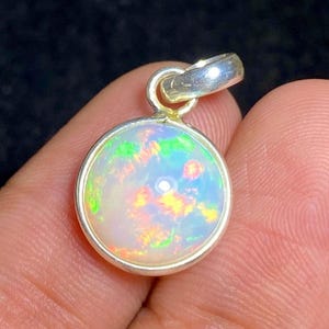 Puede incluir: Un colgante redondo con un ópalo cabujón engastado en un bisel de plata pulida. El ópalo muestra una base blanca translúcida con destellos vibrantes de color verde, naranja y amarillo. Una anilla de plata está unida a la parte superior.