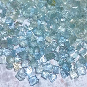 May include: A collection of raw, uncut aquamarine gemstones. The stones vary in size and shape, displaying a range of pale blue and clear hues. The rough texture and natural form of the crystals are visible.