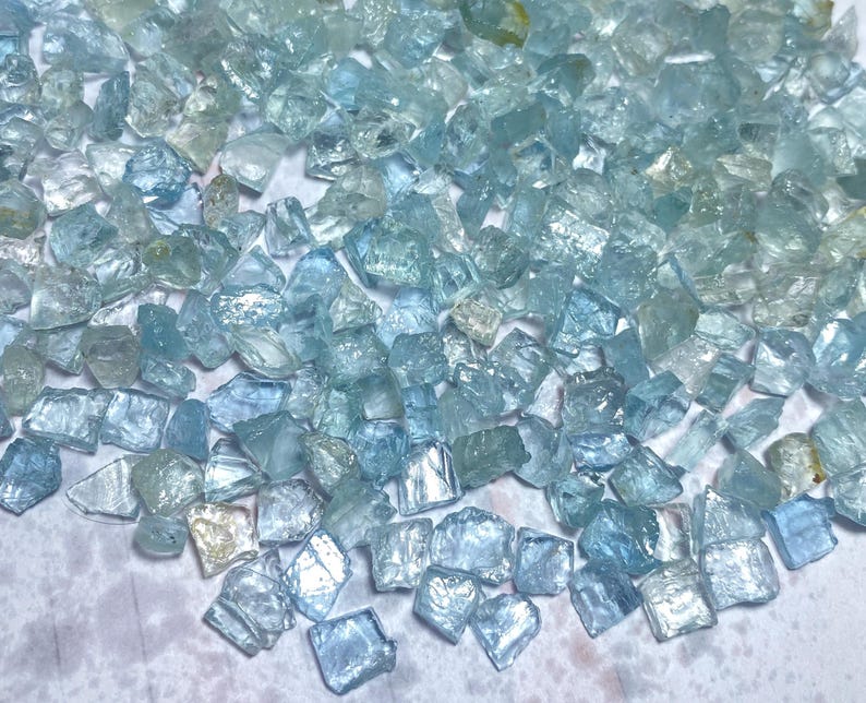 May include: A collection of raw, uncut aquamarine gemstones. The stones vary in size and shape, displaying a range of pale blue and clear hues. The rough texture and natural form of the crystals are visible.