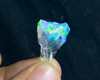 May include: A raw, iridescent opal gemstone with a fractured, irregular shape. The opal displays a vibrant play of color, including flashes of blue, green, and purple. The stone is held against a dark background, highlighting its natural beauty.