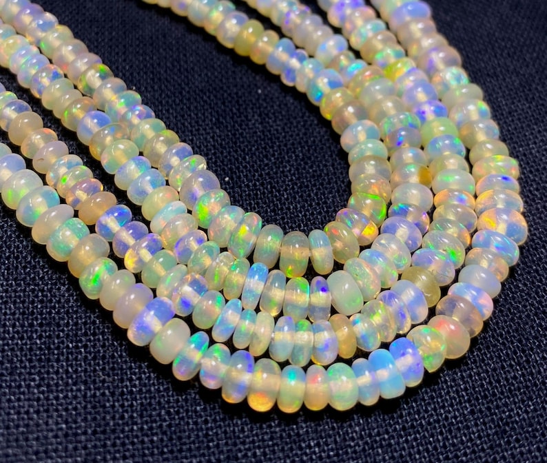 May include: Close-up of multiple strands of opal bead necklaces. The beads are a mix of white, yellow, and blue, with iridescent flashes of color. The beads are strung together, creating a layered effect. The background is a dark blue fabric.