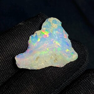 May include: A rough-cut opal gemstone with a milky white base color. The opal displays iridescent flashes of color, including green, blue, and yellow. The stone has a natural, irregular shape and a slightly rough texture.