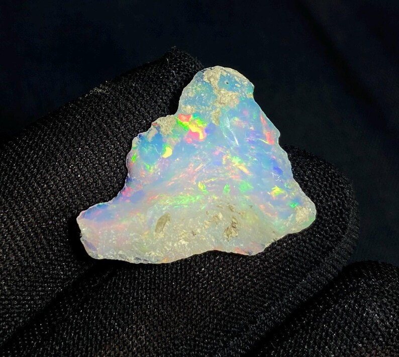 May include: A rough-cut opal gemstone with a milky white base color. The opal displays iridescent flashes of color, including green, blue, and yellow. The stone has a natural, irregular shape and a slightly rough texture.