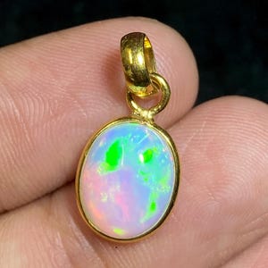 May include: An oval-shaped pendant featuring a smooth, iridescent opal gemstone set in a polished gold-toned metal bezel. The opal displays vibrant flashes of neon green, pink, and blue light against a milky white base, suspended by a gold loop.