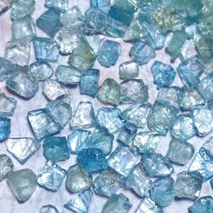 May include: A collection of raw aquamarine gemstones in various shades of blue and teal. The gemstones are irregularly shaped and appear translucent, with a sparkling, crystalline texture. The stones are scattered across a light-colored surface.