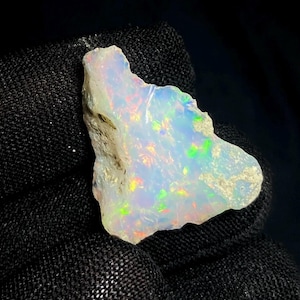 May include: A rough-cut opal gemstone with a milky white base color. The opal displays iridescent flashes of color, including green, yellow, and red. The stone is held against a black background, highlighting its natural beauty.