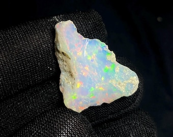 May include: A rough-cut opal gemstone with a milky white base color. The opal displays iridescent flashes of color, including green, yellow, and red. The stone is held against a black background, highlighting its natural beauty.