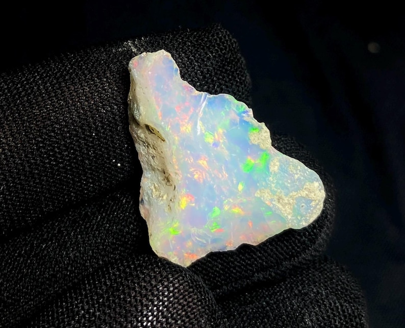 May include: A rough-cut opal gemstone with a milky white base color. The opal displays iridescent flashes of color, including green, yellow, and red. The stone is held against a black background, highlighting its natural beauty.