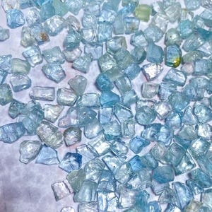 May include: A collection of raw, uncut aquamarine gemstones. The stones vary in size and shape, displaying a range of pale blue and clear hues. The gemstones are scattered across a white surface, creating a sparkling effect.