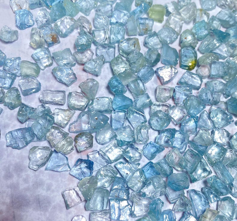 May include: A collection of raw, uncut aquamarine gemstones. The stones vary in size and shape, displaying a range of pale blue and clear hues. The gemstones are scattered across a white surface, creating a sparkling effect.