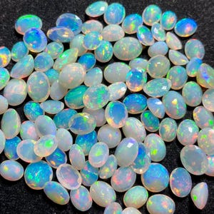 May include: A collection of oval-shaped opals in various sizes, displaying a range of colors including white, blue, green, and hints of yellow. The opals have a polished, iridescent surface, with a play of color visible throughout. The gemstones are scattered across a dark surface.