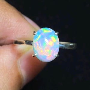 Può includere: Un anello in argento con una gemma di opale ovale a cabochon al centro. L'opale presenta una base bianca traslucida con vivaci bagliori di colore verde, giallo e arancione. La pietra è fissata da quattro griffe su una fascia metallica.
