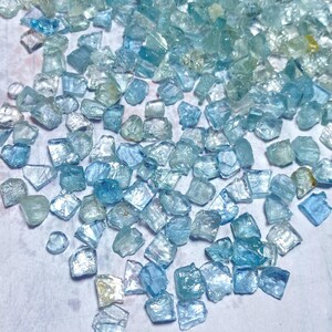 May include: A collection of raw aquamarine gemstones in various shades of blue and teal. The gemstones are irregularly shaped and appear translucent, with a slightly rough texture. The stones are scattered across a light-colored surface.