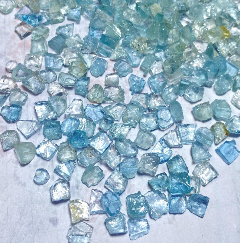 May include: A collection of raw aquamarine gemstones in various shades of blue and teal. The gemstones are irregularly shaped and appear translucent, with a slightly rough texture. The stones are scattered across a light-colored surface.