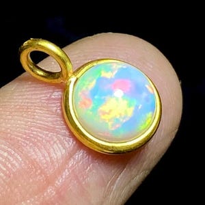 May include: A round opal pendant with a gold-colored bezel and bail. The opal displays a vibrant play of color, including blue, yellow, and pink hues. The pendant is held up by a finger against a dark background.