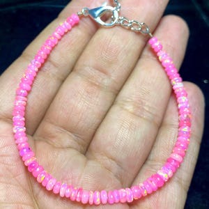 Op de afbeelding: Een armband bestaande uit kleine, ronde, felroze kralen met iriserende vlekjes. De streng is bevestigd met een zilverkleurige metalen karabijnsluiting en een verstelbare kettingverlenging, waardoor een doorlopende cirkelvormige lus van roze stenen ontstaat.