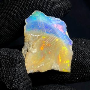 Könnte beinhalten: Ein roher Opal-Edelstein mit einer lebendigen Farbgebung. Der Stein weist eine Mischung aus Blau, Gelb und Orange auf, mit einer milchig-weißen Basis. Der Opal ist roh geschnitten und zeigt seine natürliche, unpolierte Textur. Der Edelstein wird vor dunklem Hintergrund gehalten.