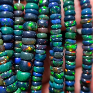 Puede incluir: Primer plano de múltiples hebras de cuentas de ópalo. Las cuentas son una mezcla de azules oscuros, verdes y marrones, con destellos de color. Las cuentas son redondas y están ensartadas, creando una exhibición texturizada y colorida.