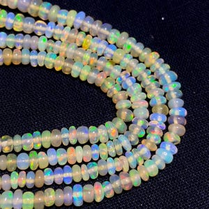 May include: Close-up of several strands of opal beads. The beads are a mix of white, yellow, green, and blue, with iridescent flashes. The beads are strung together, creating a necklace or bracelet.
