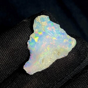 May include: A raw, uncut opal gemstone with a milky white base color. The opal displays iridescent flashes of color, including blue, green, and yellow. The stone has a rough, natural texture and irregular shape, showcasing its organic origin.