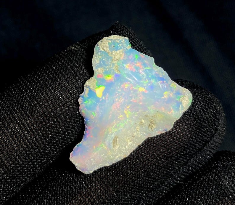 May include: A raw, uncut opal gemstone with a milky white base color. The opal displays iridescent flashes of color, including blue, green, and yellow. The stone has a rough, natural texture and irregular shape, showcasing its organic origin.