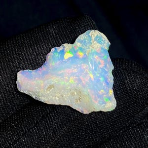 May include: A rough-cut opal gemstone with a milky white base and iridescent flashes of color. The stone displays a play of color with hues of green, yellow, and blue. The opal is set against a dark, textured background.