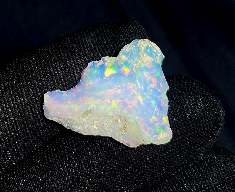 May include: A rough-cut opal gemstone with a milky white base and iridescent flashes of color. The stone displays a play of color with hues of green, yellow, and blue. The opal is set against a dark, textured background.