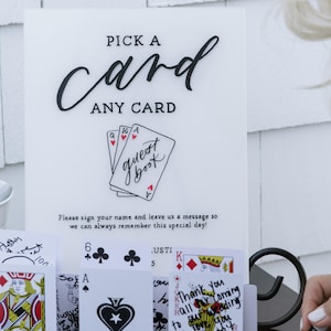 May include: A white sign with the words "PICK A card ANY CARD" in black script. Below, a drawing of playing cards with the words "guest book". Cards with messages are displayed below.