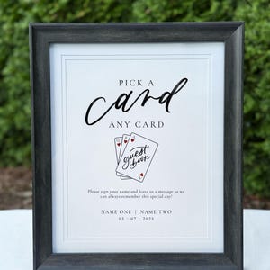 May include: A framed sign with the text "PICK A card ANY CARD" in black script. Below is a graphic of playing cards with the words "guest book." The sign includes space for names and a date, perfect for a wedding or event.