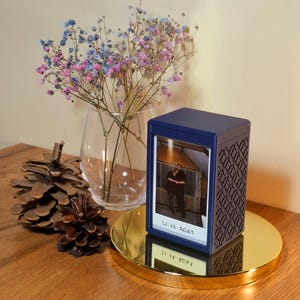 May include: A navy blue box with a decorative pattern and a photo of a person, dated 11.12.2023. The box is on a gold-coloured circular tray. A clear glass vase with colourful flowers and pine cones are also present on a wooden surface.