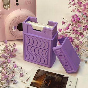 Op de afbeelding: Een lila fotodoos met een golfpatroon, met witte foto's. Een bijpassend deksel ligt ernaast open. Een roze Instax Mini 12 camera staat op de achtergrond, samen met foto's en takjes paarse bloemen.